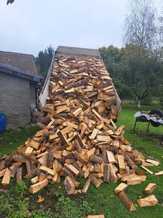 Ciężarówka z wywrotką wysypująca stos pociętego drewna na trawnik, w tle budynki i drzewa.