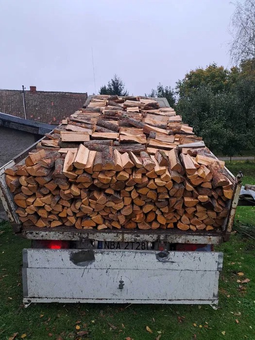 Ciężarówka z otwartą skrzynią załadowana po brzegi pociętym drewnem kominkowym, widok z tyłu, tablica rejestracyjna widoczna, w tle budynki i drzewa