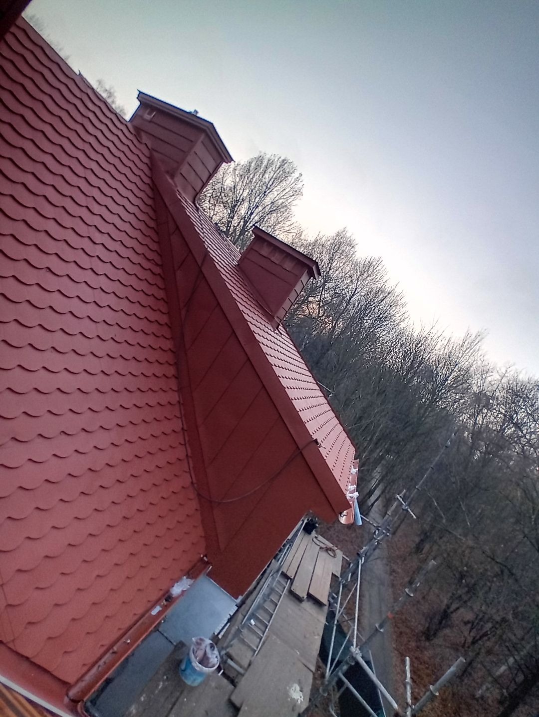 Czerwony dach z dachówki karpiówki, widok z góry, z kominami i rusztowaniem. Tło stanowią drzewa bez liści i szare niebo. Nietypowy kąt ujęcia.