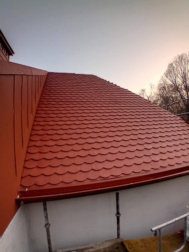 Czerwony dach z dachówki karpiówki, widok z dołu pod kątem. Widoczne orynnowanie i fragment ściany budynku. W tle drzewa bez liści i jasne niebo.