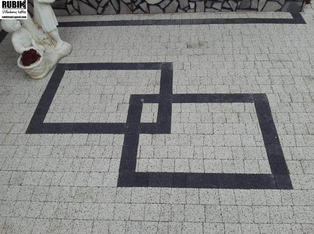 Ułożona kostka brukowa w jasnych odcieniach z wkomponowanym wzorem dwóch przenikających się czarnych kwadratów. Dekoracyjna figurka z boku.