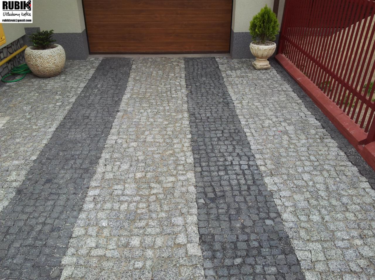 Granitowa kostka w pasy prowadzi do garażu. Ciemnoszare i jasnoszare kostki tworzą wzór. Po bokach donice z roślinami i czerwone ogrodzenie.