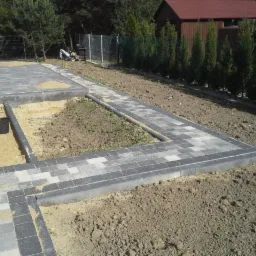 Świeżo ułożona, szara kostka brukowa tworząca ścieżkę i obramowanie rabaty w ogrodzie. W tle widoczny dom z czerwonym dachem i tuje.