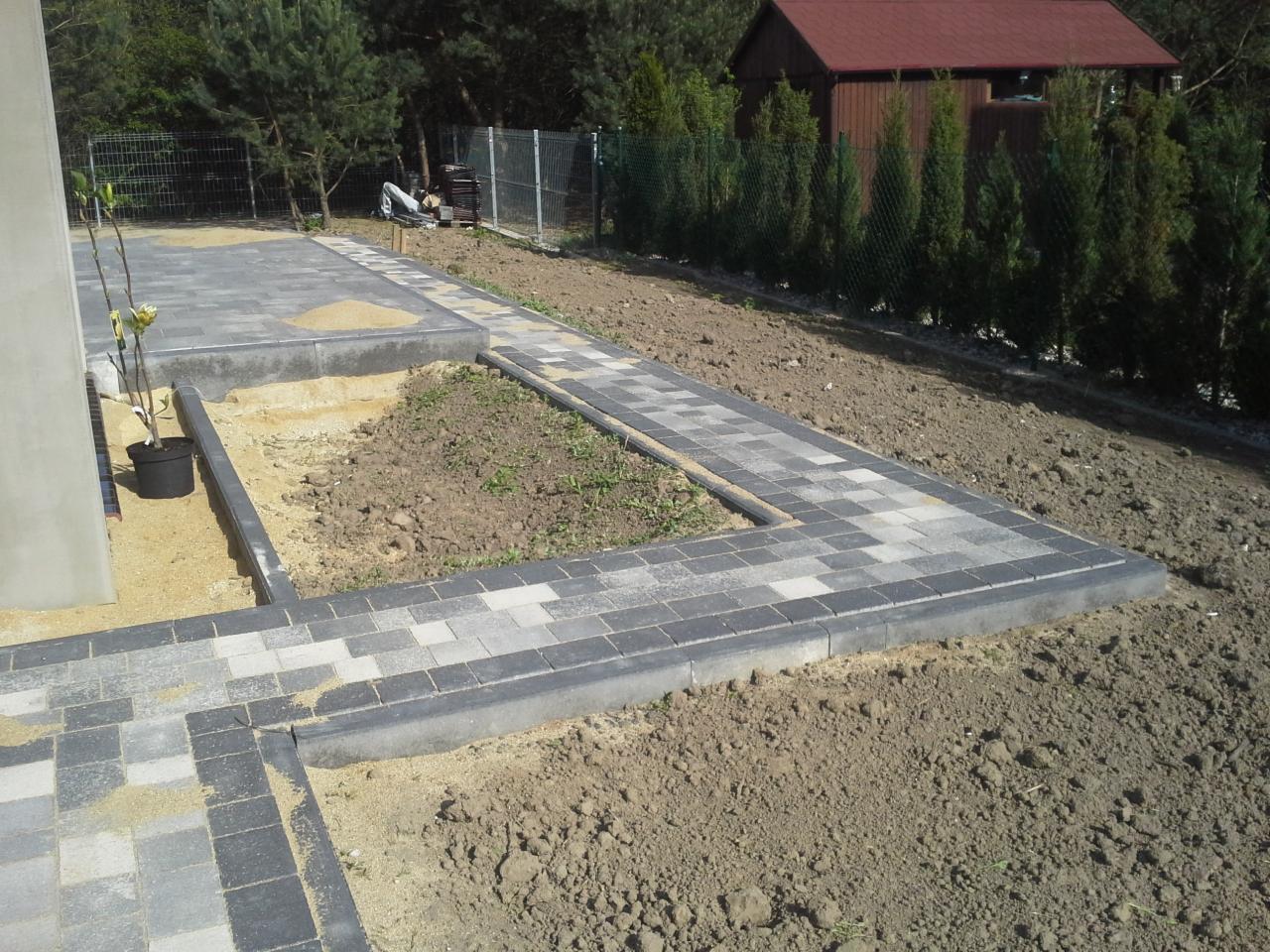Świeżo ułożona, szara kostka brukowa tworząca ścieżkę i obramowanie rabaty w ogrodzie. W tle widoczny dom z czerwonym dachem i tuje.