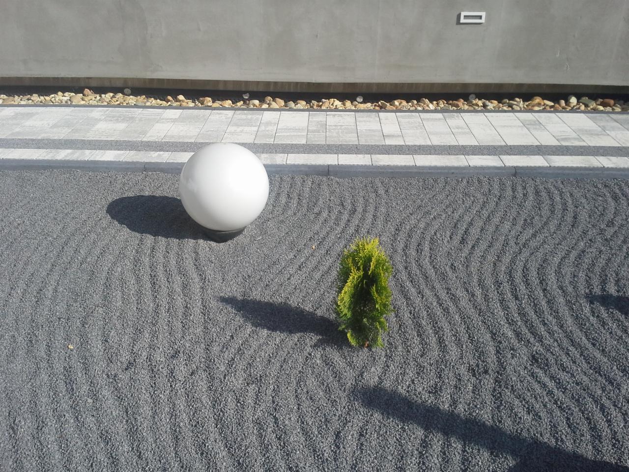 Ogród zen z szarego żwiru z wygrabionymi falami, białą kulą i małym drzewkiem iglastym. W tle betonowy mur z oświetleniem i pasem małych kamieni.