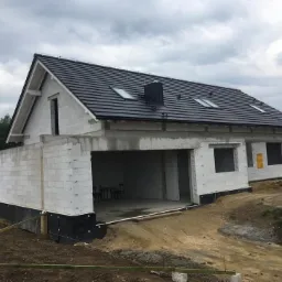 Budynek jednorodzinny w trakcie budowy: surowy stan, widoczna czarna hydroizolacja fundamentów, białe ściany z betonu komórkowego, czarny dach z oknami dachowymi, teren wokół w stanie przygotowania.