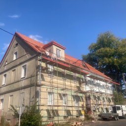 Firma Remontowo Budowlana - Dekarstwo Rafał Cichy - Prace na dachu budynku wielorodzinnego, widoczne rusztowania, częściowo nowy czerwony dach, pracownicy na dachu, biały bus firmy budowlanej na ulicy.