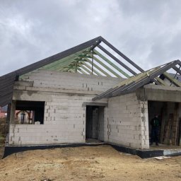 Firma Remontowo Budowlana - Dekarstwo Rafał Cichy - Surowy stan domu jednorodzinnego w trakcie budowy, widoczna konstrukcja drewniana dachu, ściany z białego bloczka, ziemia wokół fundamentów, pracownik w środku budynku.