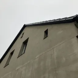Widok z dołu na szczyt budynku z nowym, czarnym pokryciem dachu i orynnowaniem, na tle pochmurnego nieba. Widoczne trzy okna o nietypowych, wydłużonych kształtach.