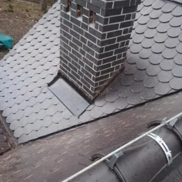 Dach pokryty gontem bitumicznym w kształcie rybiej łuski, z kominem z ciemnej cegły z jasnymi spoinami; widoczne orynnowanie z metalowymi uchwytami.