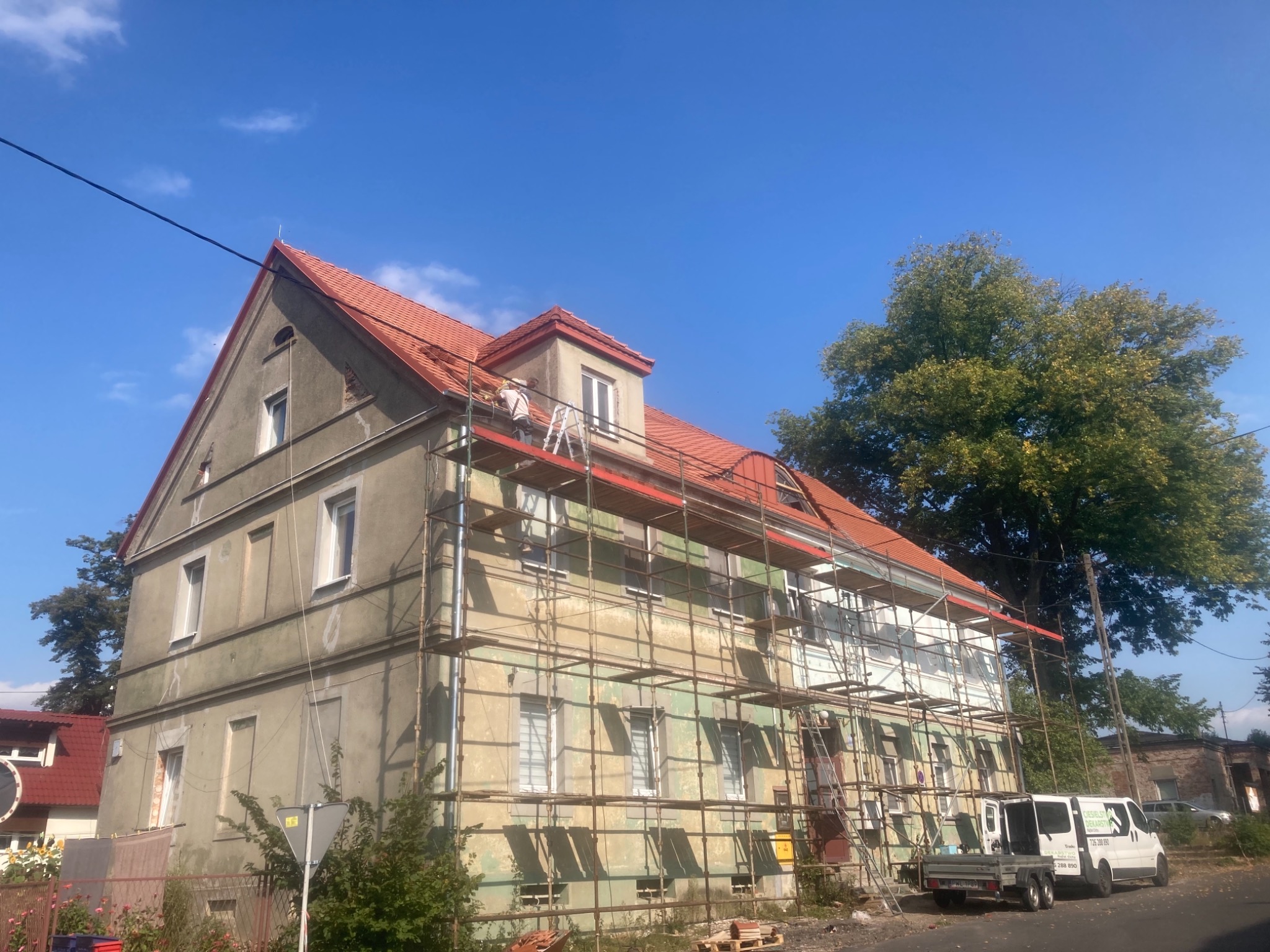 Prace na dachu budynku wielorodzinnego, widoczne rusztowania, częściowo nowy czerwony dach, pracownicy na dachu, biały bus firmy budowlanej na ulicy.