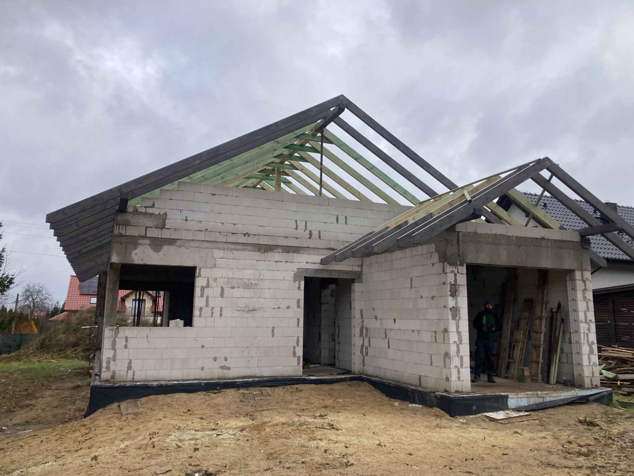 Surowy stan domu jednorodzinnego w trakcie budowy, widoczna konstrukcja drewniana dachu, ściany z białego bloczka, ziemia wokół fundamentów, pracownik w środku budynku.