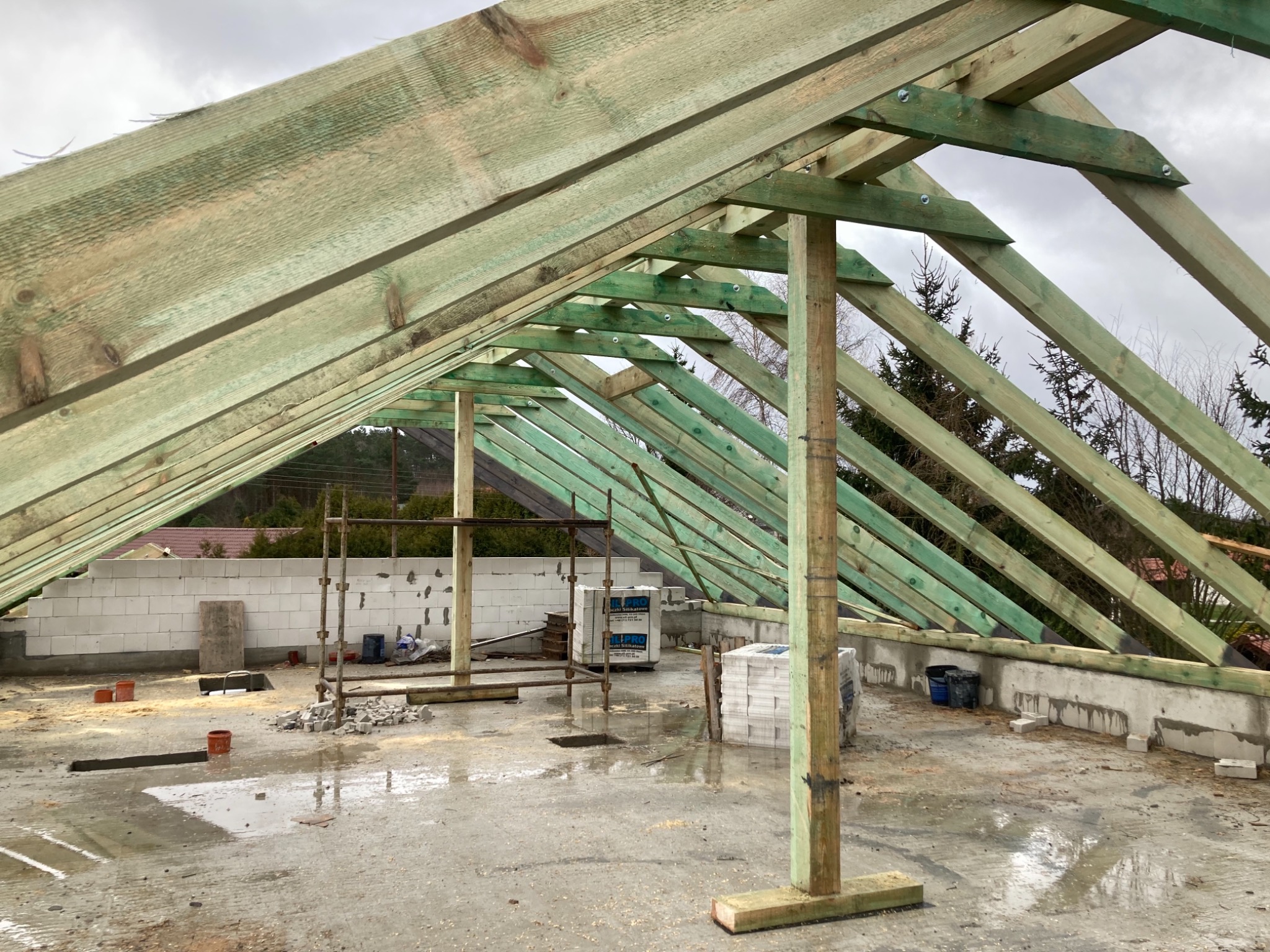 Surowa, impregnowana konstrukcja drewniana dachu z widocznymi elementami mocującymi, w trakcie budowy, z paletami bloczków silikatowych na mokrym podłożu i rusztowaniami w tle.