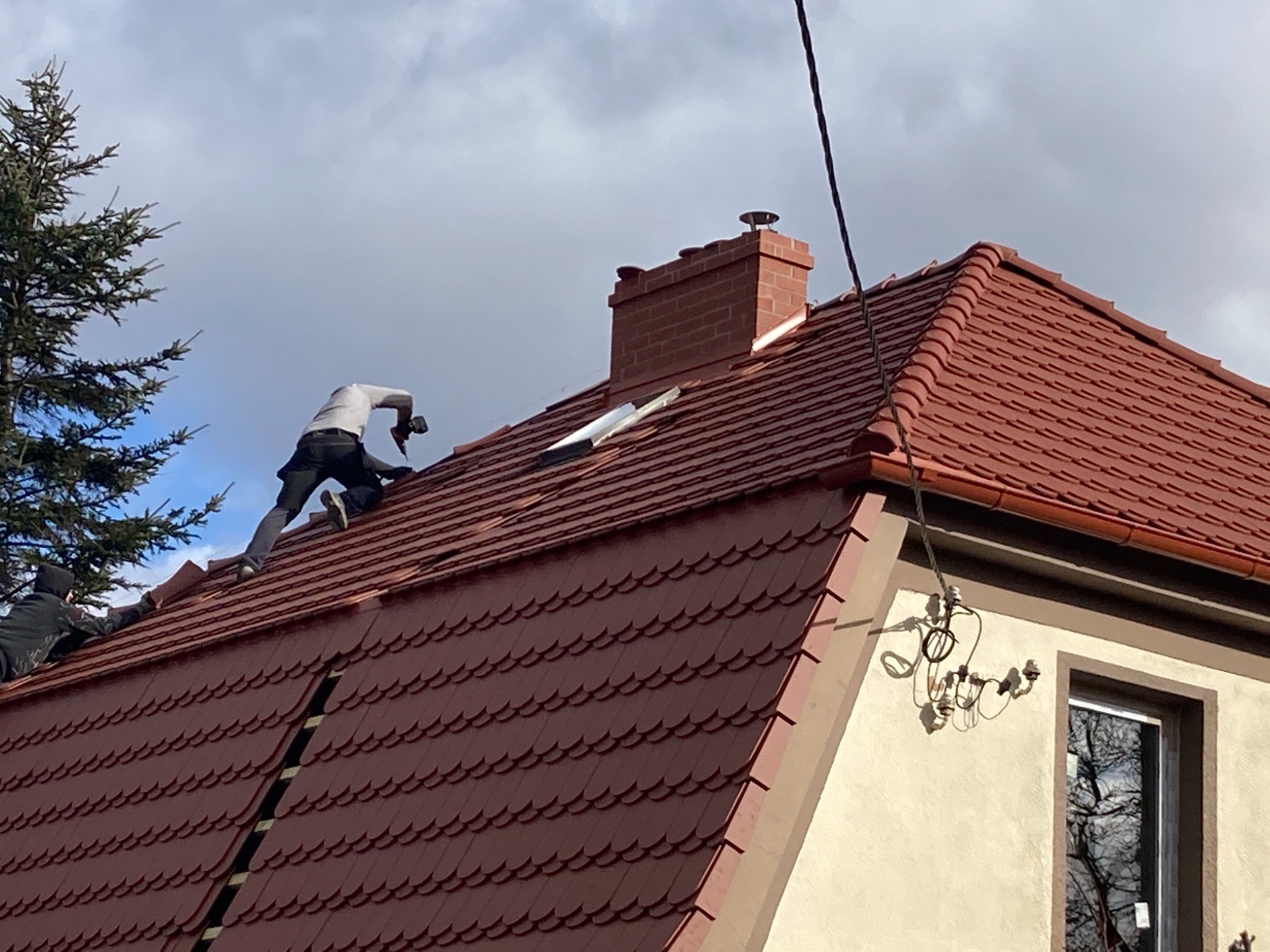 Dekarze montują czerwoną dachówkę na skośnym dachu domu jednorodzinnego z ceglanym kominem, widoczne narzędzia i fragment instalacji elektrycznej na ścianie.
