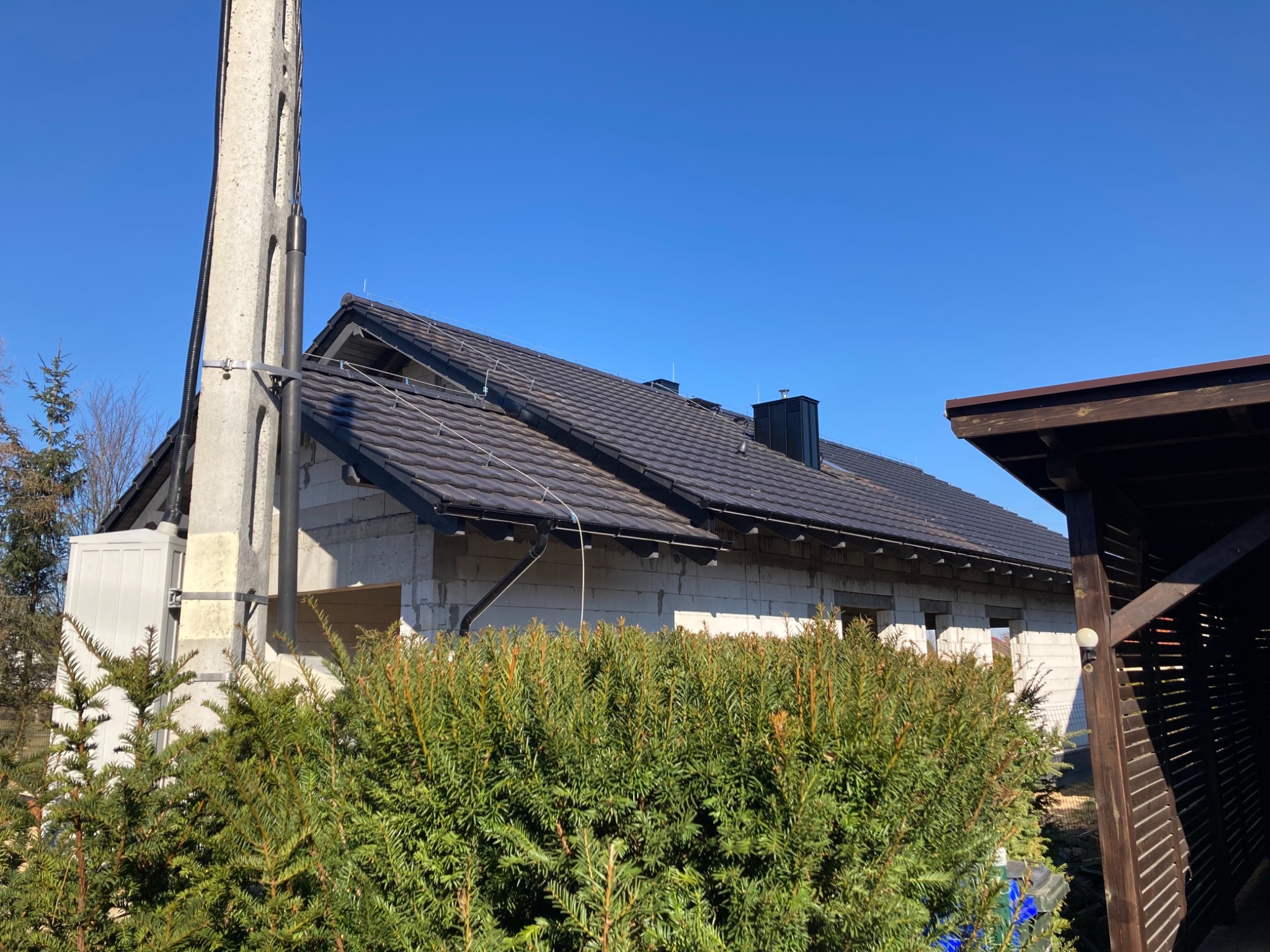 Surowy stan budynku z ciemną dachówką na jasnym niebie, widoczna betonowa podpora energetyczna i fragment drewnianej konstrukcji z boku, zieleń krzewów na pierwszym planie.