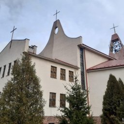 Quoss Agnieszka Sidor - Kościół o nietypowej architekturze z trzema krzyżami, białymi ścianami z fakturą i brązowymi ramami okiennymi, częściowo zasłonięty przez zielone drzewa iglaste, sfotografowany w pochmurny dzień.