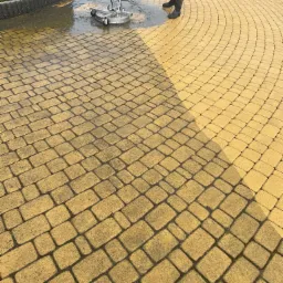 Żółta kostka brukowa czyszczona myjką ciśnieniową z dyszą obrotową, widoczne różnice w kolorze między umytą a brudną powierzchnią, osoba w gumowych butach obsługuje urządzenie.