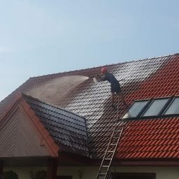 Quoss Agnieszka Sidor - Częściowo oczyszczony dach z czerwonej dachówki, pracownik na drabinie czyści go myjką ciśnieniową, widoczne okna dachowe.