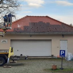 Quoss Agnieszka Sidor - Pracownicy na podnośniku koszowym czyszczą dach z czerwonej dachówki, widoczna wyraźna różnica między umytą a brudną częścią dachu, na dole garaż z bramą i znak parkingowy.