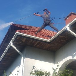 Quoss Agnieszka Sidor - Mężczyzna w uprzęży, stojąc na dachu z czerwonej dachówki, czyści go za pomocą myjki ciśnieniowej, strumień wody spływa po krawędzi dachu do rynny, błękitne niebo w tle.