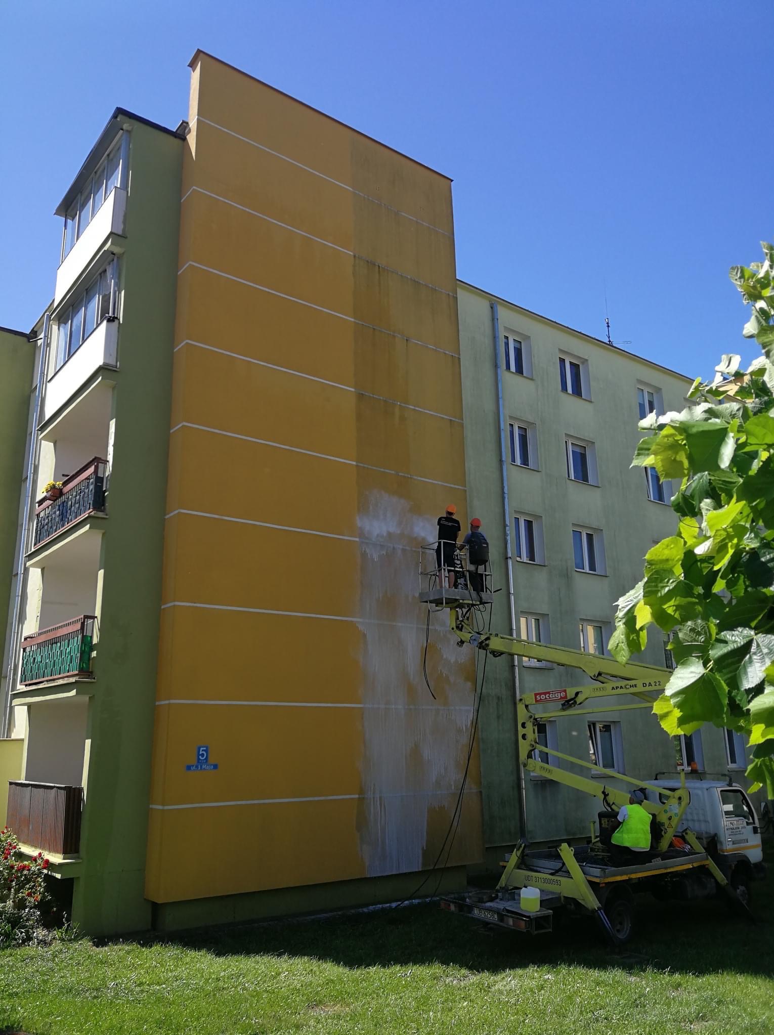 Dwaj pracownicy na podnośniku koszowym czyszczą fragment żółtej elewacji budynku, widoczne różnice w kolorze przed i po czyszczeniu, znak z numerem 5 na ścianie.