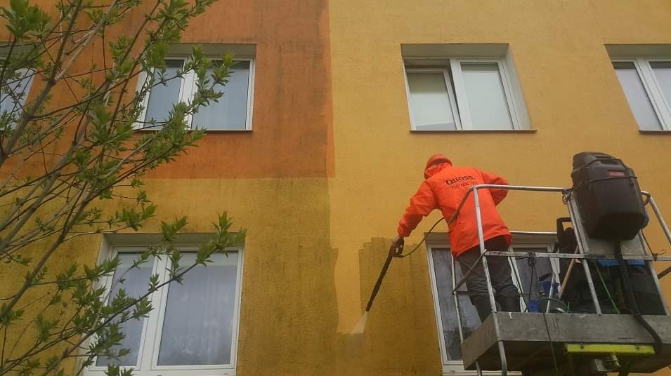 Pracownik w pomarańczowym kombinezonie na podnośniku koszowym czyści elewację budynku, widoczny efekt różnicy między umytą a brudną powierzchnią, z lewej strony kadru gałęzie drzewa.