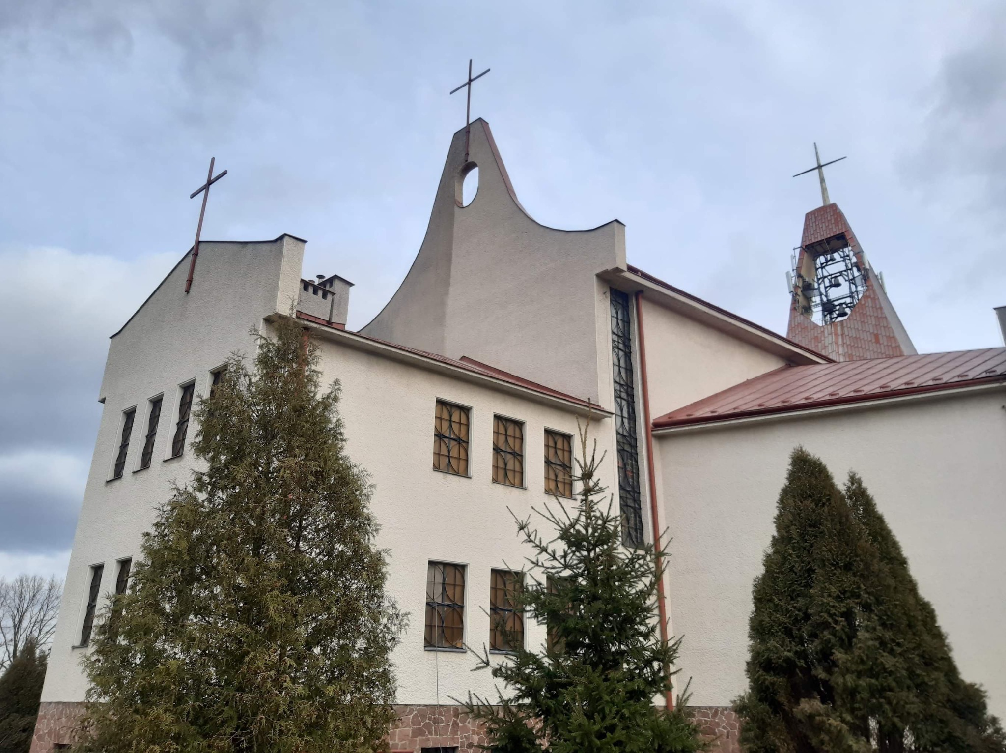 Kościół o nietypowej architekturze z trzema krzyżami, białymi ścianami z fakturą i brązowymi ramami okiennymi, częściowo zasłonięty przez zielone drzewa iglaste, sfotografowany w pochmurny dzień.