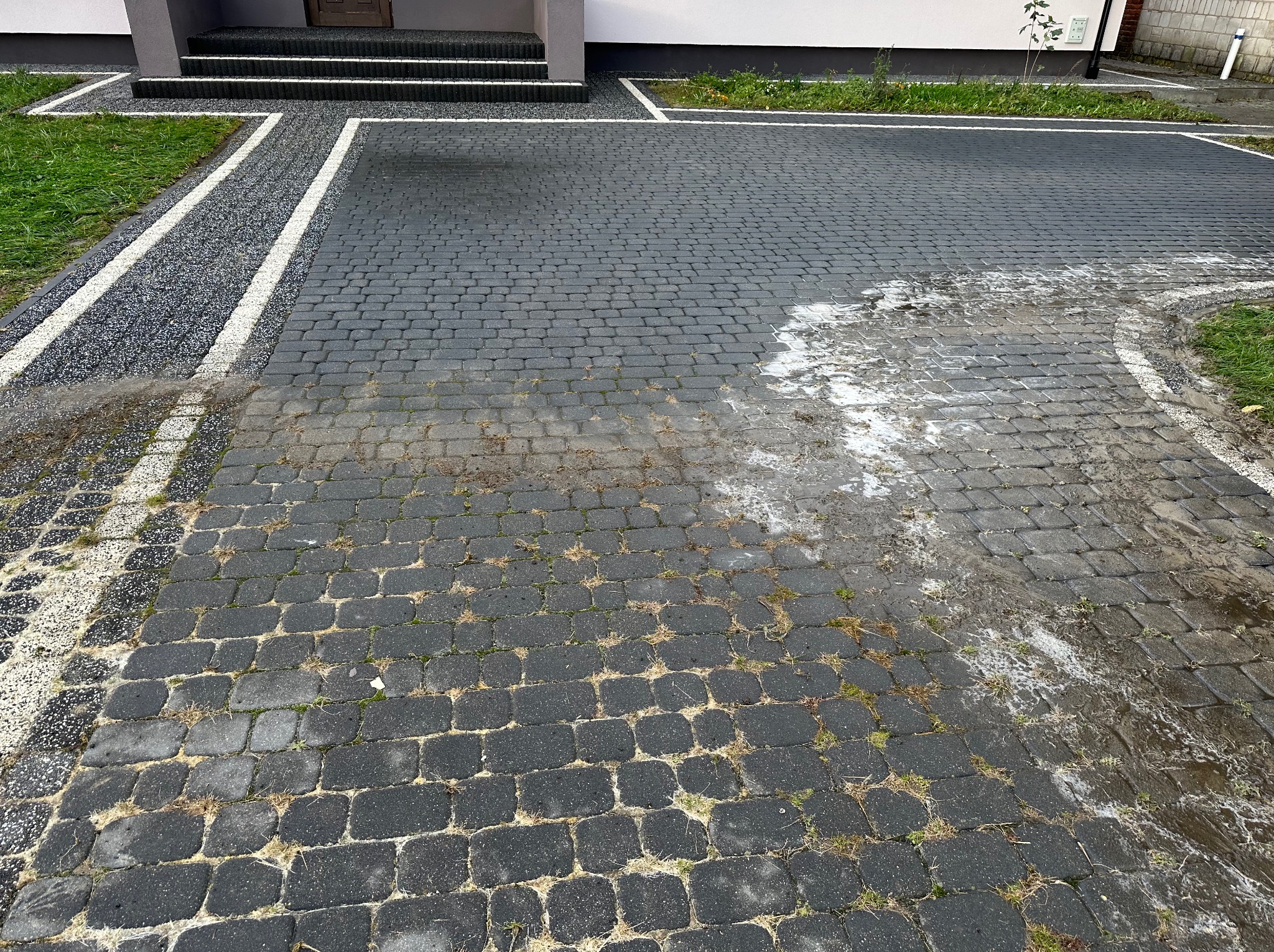 Częściowo umyta, czarna kostka brukowa przed domem w Wojciechowie, z widocznymi smugami wody i resztkami piany po czyszczeniu, kontrastująca z suchą częścią nawierzchni.