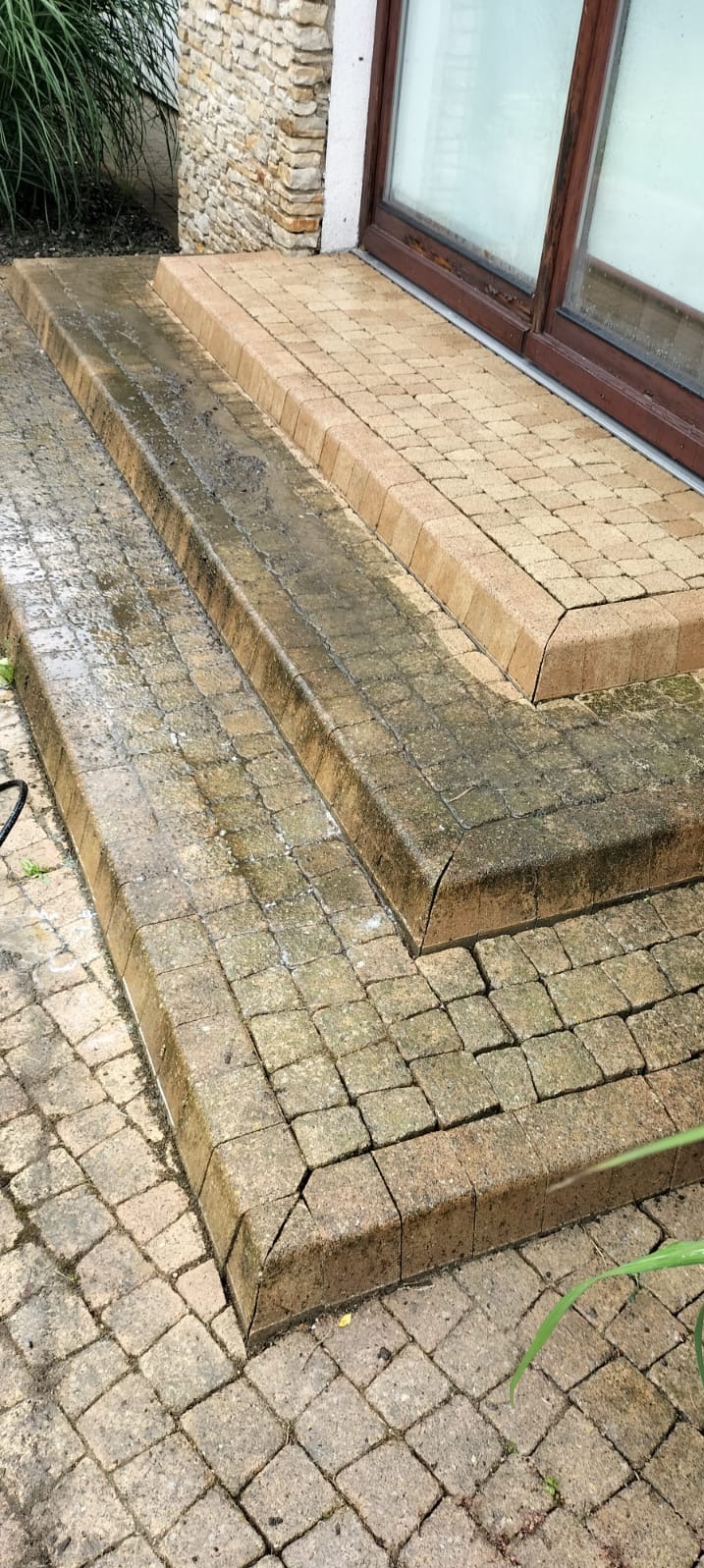 Schody z kostki brukowej częściowo umyte, widoczna różnica w kolorze między czystą a brudną powierzchnią, zarys okna w tle.