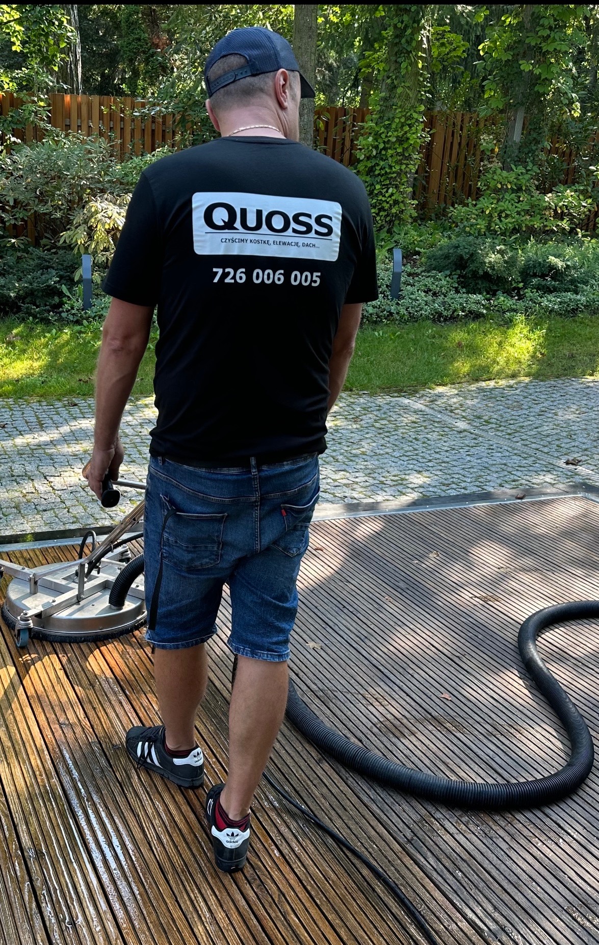 Pracownik w czarnej koszulce z logo firmy QUOSS, czyszczący drewniany taras za pomocą okrągłej myjki ciśnieniowej; widoczny wąż doprowadzający wodę i mokra powierzchnia po czyszczeniu, w tle...