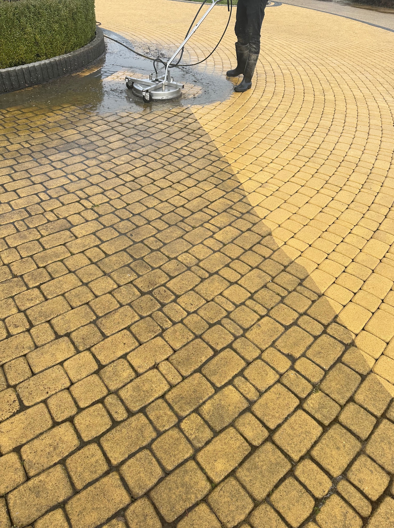 Żółta kostka brukowa czyszczona myjką ciśnieniową z dyszą obrotową, widoczne różnice w kolorze między umytą a brudną powierzchnią, osoba w gumowych butach obsługuje urządzenie.
