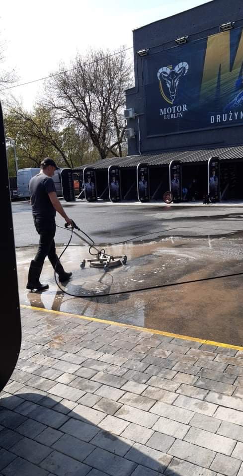 Pracownik w czarnym ubraniu i gumowych butach czyści betonową nawierzchnię myjką ciśnieniową z rotacyjną dyszą, na tle budynku z reklamą Motor Lublin.