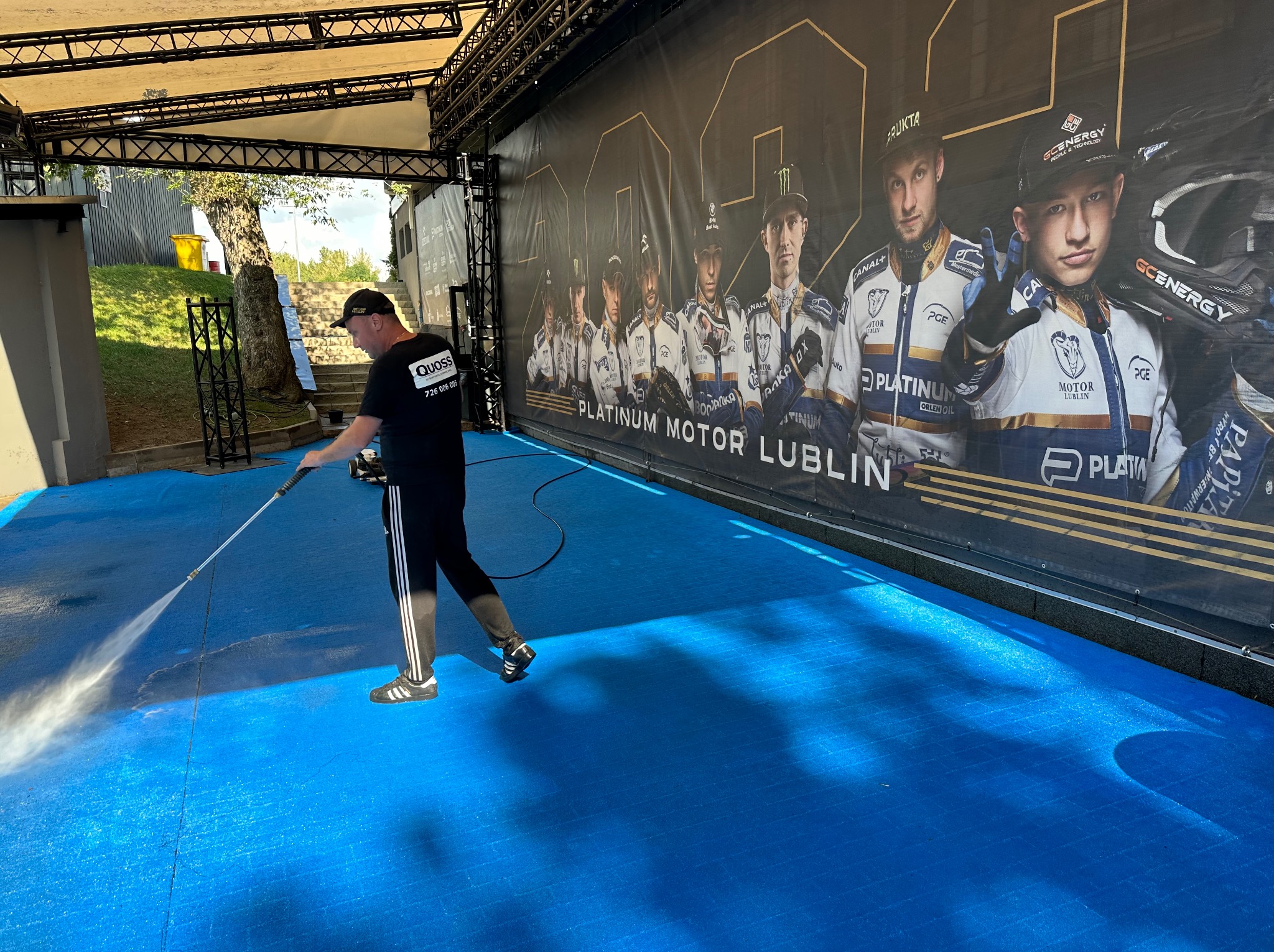 Pracownik w czarnym uniformie z logo firmy czyści niebieską nawierzchnię pod zadaszeniem za pomocą myjki ciśnieniowej. W tle widoczny baner reklamowy Platinum Motor Lublin.