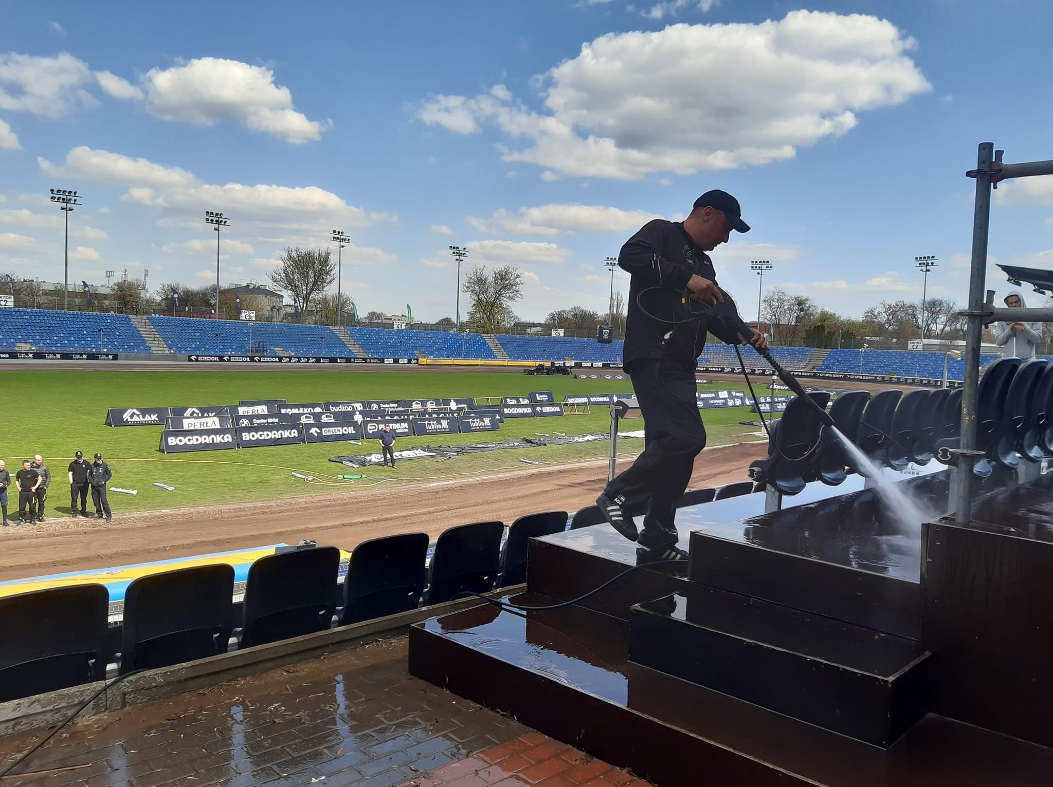Pracownik w czarnym kombinezonie i czapce czyści trybuny stadionu sportowego za pomocą myjki ciśnieniowej, widoczne mokre powierzchnie i woda spływająca po schodach, w tle niebieskie krzesełka...