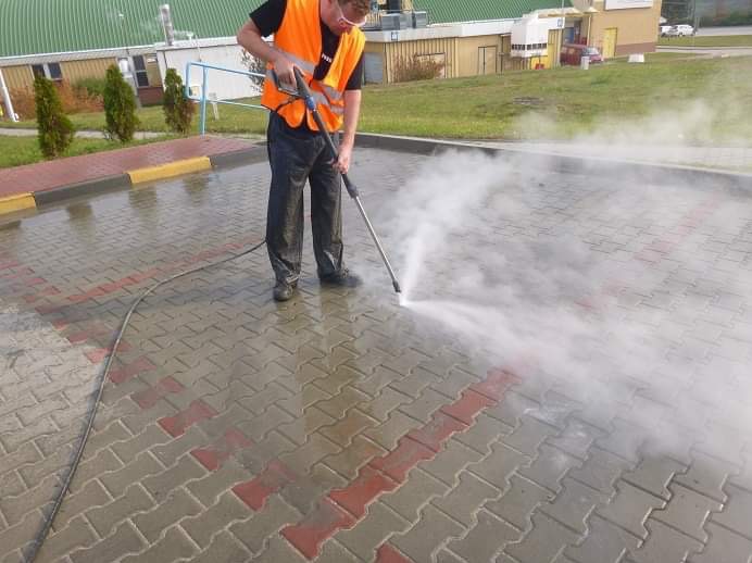 Pracownik w pomarańczowej kamizelce odblaskowej czyści kostkę brukową za pomocą myjki ciśnieniowej, strumień wody unosi się w postaci pary, widoczne mokre i czyste fragmenty kostki. Częściowo...
