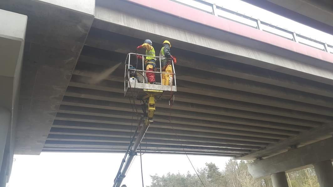 Pracownicy na podnośniku koszowym czyszczą konstrukcję betonową mostu metodą strumieniową pod ciśnieniem, w tle widoczny las.