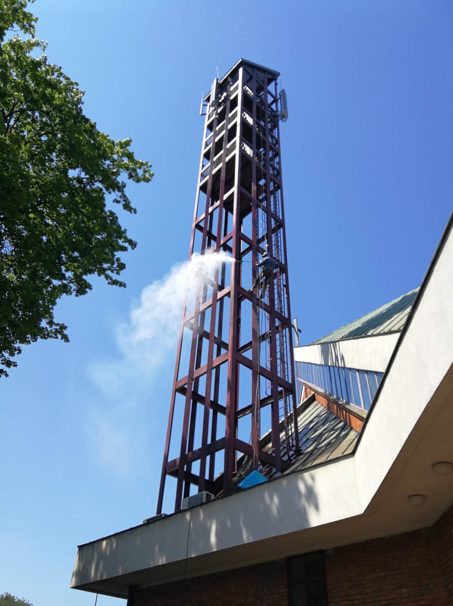 Pracownik na wysokości myje wodą pod ciśnieniem konstrukcję dzwonnicy kościoła, widoczna część dachu budynku i błękitne niebo.