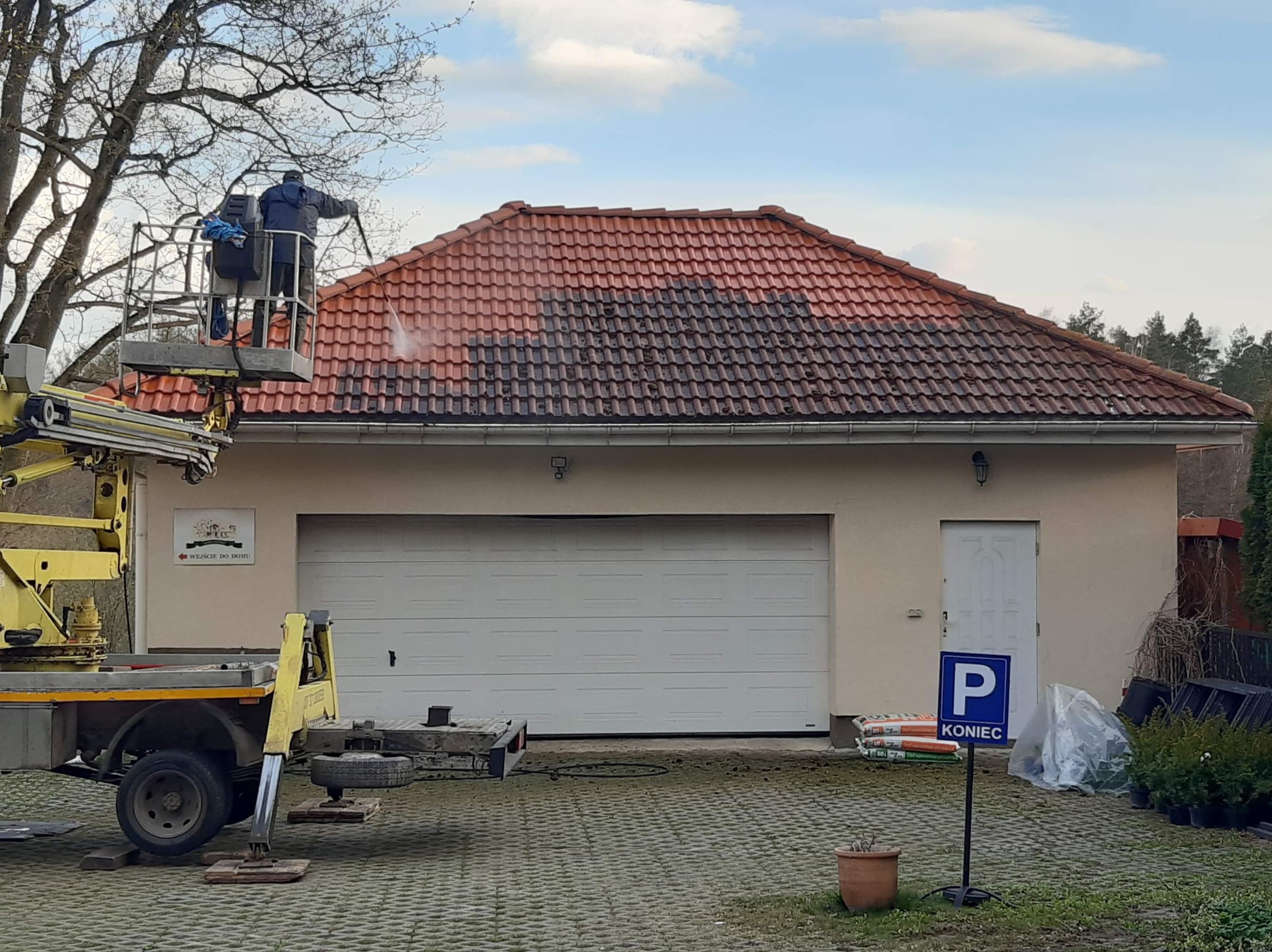 Pracownicy na podnośniku koszowym czyszczą dach z czerwonej dachówki, widoczna wyraźna różnica między umytą a brudną częścią dachu, na dole garaż z bramą i znak parkingowy.
