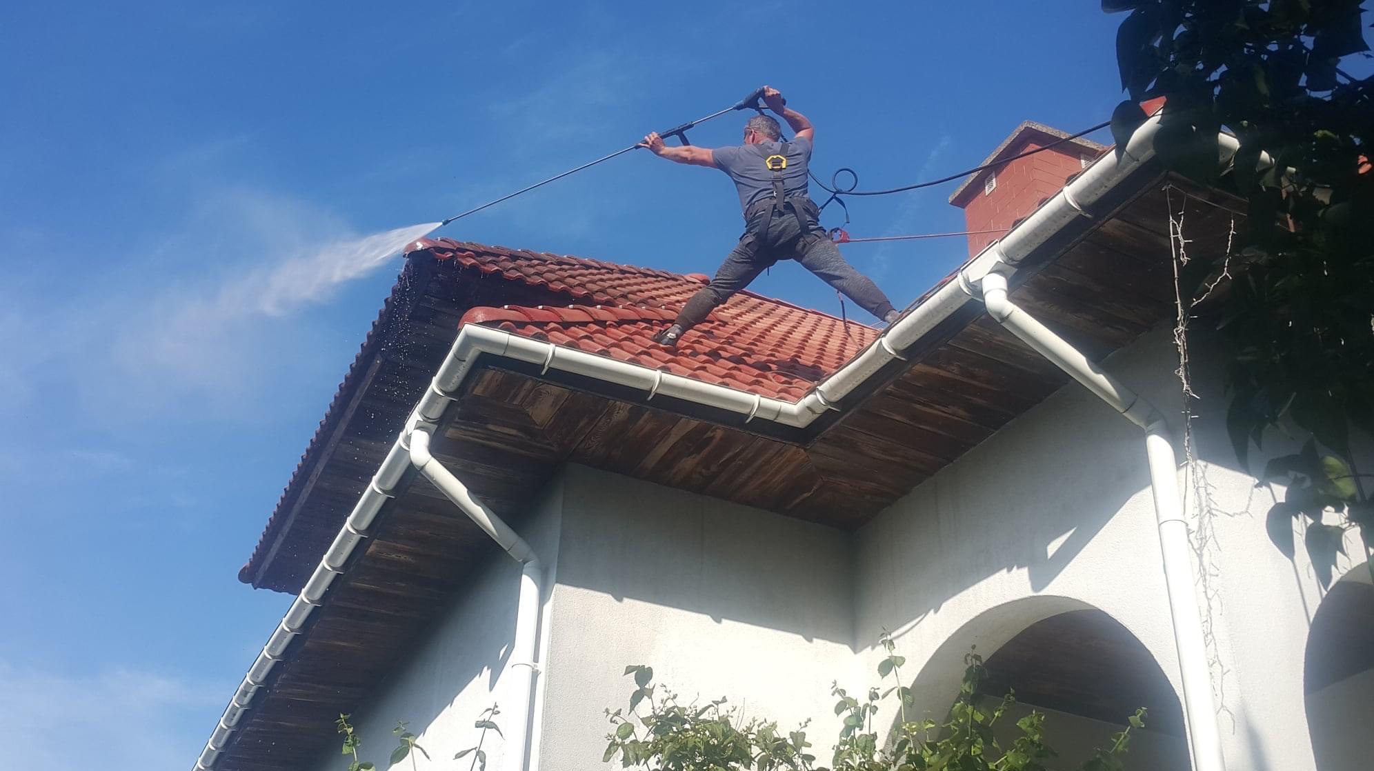 Mężczyzna w uprzęży, stojąc na dachu z czerwonej dachówki, czyści go za pomocą myjki ciśnieniowej, strumień wody spływa po krawędzi dachu do rynny, błękitne niebo w tle.