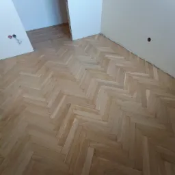 Ułożony parkiet w jodełkę, jasne drewno, widoczne ślady po listwach przypodłogowych, surowe ściany w trakcie wykończenia, gniazdka elektryczne bez osłon.