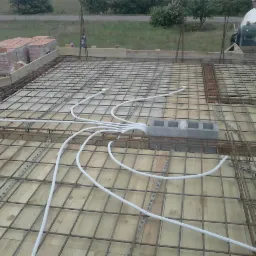 Zbrojenie płyty fundamentowej z rozprowadzonymi rurami instalacji elektrycznej, widok z góry. Na obrzeżach ułożone cegły i bloczki betonowe, w tle zieleń i betoniarka.