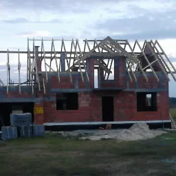 Surowy stan domu z czerwonej cegły z drewnianą konstrukcją dachu na tle pochmurnego nieba. Widoczne pustaki i piasek. Etap budowy domu jednorodzinnego.