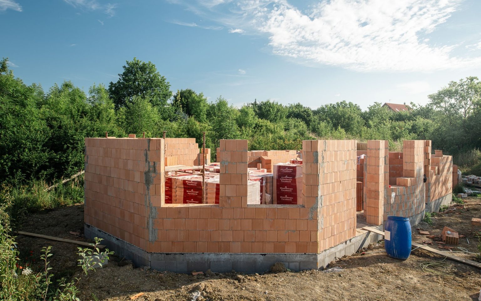 Wznoszenie ścian parteru domu z czerwonej cegły Porotherm, widoczne palety z cegłami wewnątrz budynku, błękitne niebo i zieleń drzew w tle.