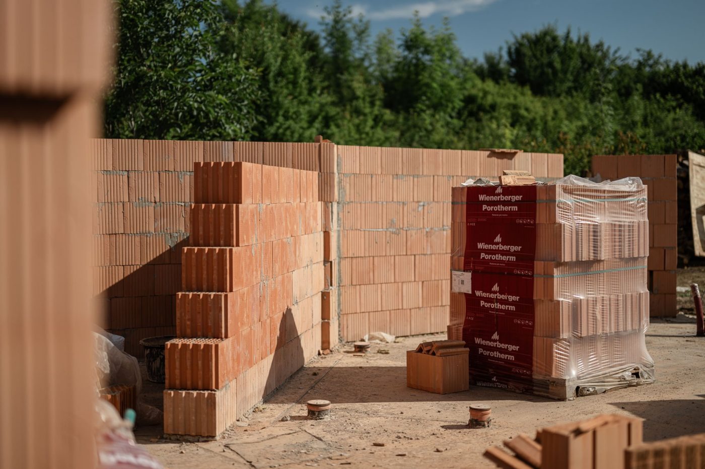 Budowa ściany z czerwonych bloczków Porotherm, paleta z zapasem materiału na placu budowy. Widoczne fragmenty muru i zielone drzewa w tle.