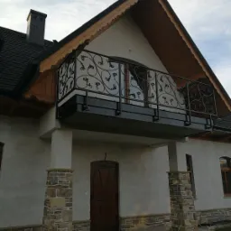 Elewacja domu z balkonem z ozdobną, metalową balustradą z motywem roślinnym, wykończona kamiennymi elementami dekoracyjnymi wokół kolumn i u podstawy ścian.