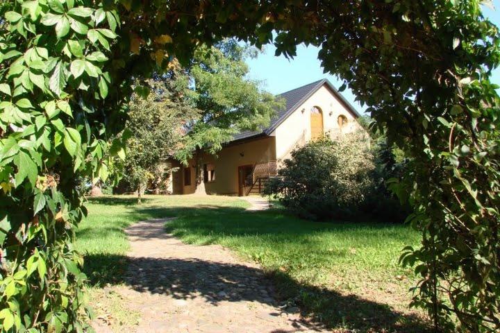 Malowniczy widok na parterowy dom z poddaszem, widziany przez zieloną, naturalną bramę z pnących roślin. Uroku dodaje kamienista ścieżka prowadząca do wejścia.