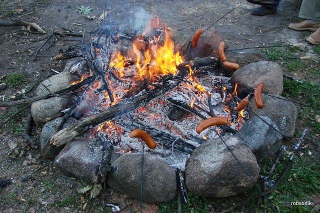 Ognisko otoczone kamieniami, na których pieką się kiełbaski. W tle widoczne nogi osób uczestniczących w ognisku. Widok z góry na palenisko.