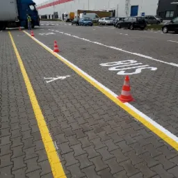 Oznakowane szarą kostką brukową miejsca parkingowe dla autobusów i pieszych przed budynkiem magazynowym z użyciem żółtej i białej farby oraz pomarańczowo-białych pachołków.