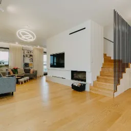 Jasne, przestronne wnętrze salonu z drewnianą podłogą, schodami z czarną balustradą, kominkiem w zabudowie, szarą sofą i okrągłym, nowoczesnym żyrandolem.