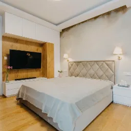 Nowoczesna sypialnia z drewnianą ścianą za telewizorem, tapicerowanym zagłówkiem w romby, parkietem i widokiem na łazienkę z umywalką.